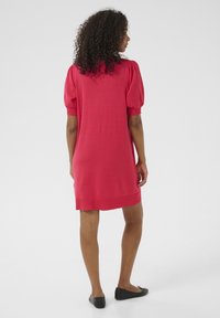 Rode gebreide jurk met korte pofmouwen, knie-lengte, ribbelzoom, zachte textuur, gecombineerd met zwarte flats. Achteraanzicht tegen een lichte achtergrond.