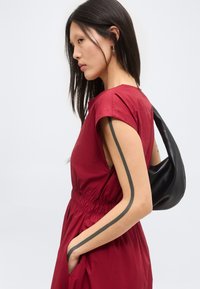 Γυναίκα με μακριά μαύρα μαλλιά φοράει αμάνικο κόκκινο φόρεμα και κρατά μαύρη τσάντα ώμου, δείχνοντας ένα μαύρο κάθετο τατουάζ στο χέρι.