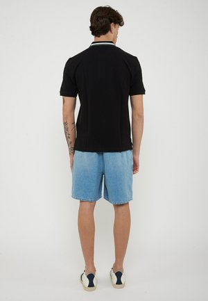 Uomo visto di spalle che indossa una polo nera a maniche corte, pantaloncini di jeans azzurri, scarpe da ginnastica bianche e un tatuaggio floreale sull'avambraccio sinistro.