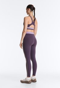 Femme aux cheveux tressés portant un soutien-gorge de sport violet avec une découpe et un legging assorti, debout de profil sur fond blanc.