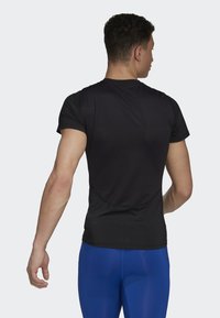 Camiseta deportiva negra de manga corta, con un escote redondeado y una textura suave, llevada con leggings ajustados azules. Vista trasera del modelo.