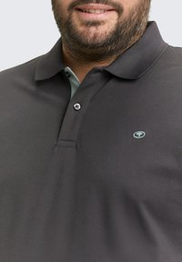 Brodaty mężczyzna w ciemnoszarej koszulce polo z jasnoszarym wewnętrznym kołnierzykiem i małym haftowanym logo na piersi.