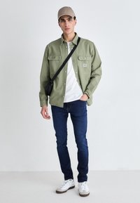 Light green button-up shirt over a white T-shirt, blue jeans, white sneakers, and a beige cap; black crossbody bag.