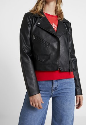 Žena oblečená v černé kožené motoristické bundě se stříbrnými zipy, pod ní červená košile a modré džíny, stojící před jednoduchým pozadím.