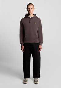 Hættebluse i mørkegrå med en frontlomme og gult logo. Båret med sorte, løstsiddende bukser og lyse sneakers.
