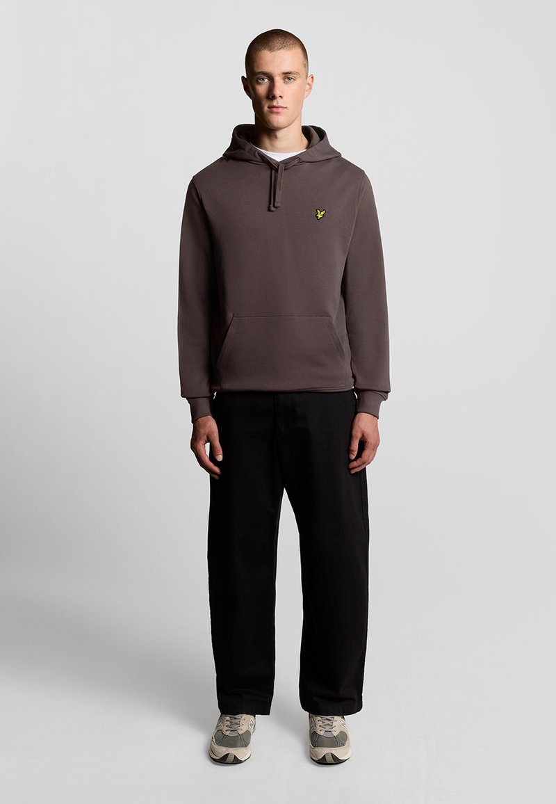 Felpa con cappuccio grigia scura con tasca frontale e logo giallo. Indossata con pantaloni neri larghi e sneakers chiare.