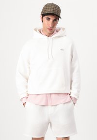 Felpa bianca con cappuccio e tasca a marsupio, con logo ricamato, sovrapposta a una maglietta a righe rosa e abbinata a pantaloni corti bianchi testurizzati.