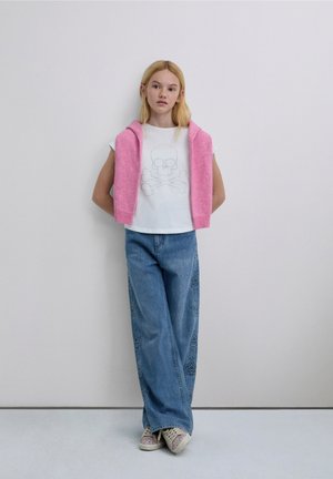 White t-shirt with skull and crossbones design, pink knit wrap around shoulders, blue wide-leg jeans with star patterns, beige sneakers.