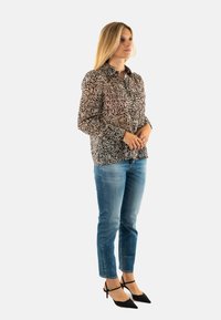 Blusa a maniche lunghe, a taglio morbido, in tessuto trasparente con un motivo a foglie nero e beige; abbinata a jeans azzurri chiari e tacchi a punta neri.