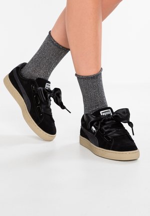 Nogi w szarych prążkowanych skarpetach oraz czarne sneakersy Puma z satynowymi sznurowadłami i beżowymi podeszwami na białym tle.