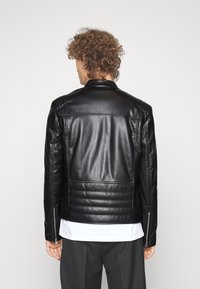 Veste en cuir noire avec une coupe ajustée, des accents matelassés aux épaules et des poignets zippés. Texture lisse avec un style épuré et minimaliste.