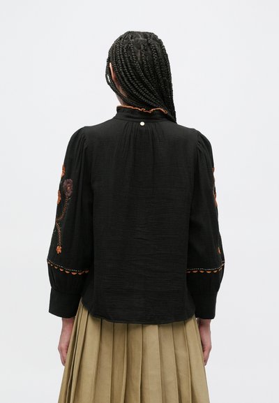 Črna bluza iz teksturirane tkanine z vezenimi rdečimi in oranžnimi cvetovi na rokavih ter z gumbi na ovratniku.