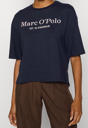 Persoon die een marineblauw Marc O'Polo t-shirt draagt met de tekst "EST. IN STOCKHOLM" en bruine geplooide broek, staand tegen een lichte achtergrond.