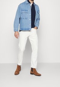 Džínová bunda ve světle modré barvě s dvěma náprsními kapsami, tmavě modré polo triko pod tím, bílé slim-fit kalhoty a hnědé semišové kotníkové boty.