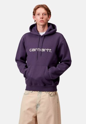 Felpa viola con tasca frontale, polsini e orlo a costine, con il logo "Carhartt" bianco. Abbinata a pantaloni larghi di colore chiaro.