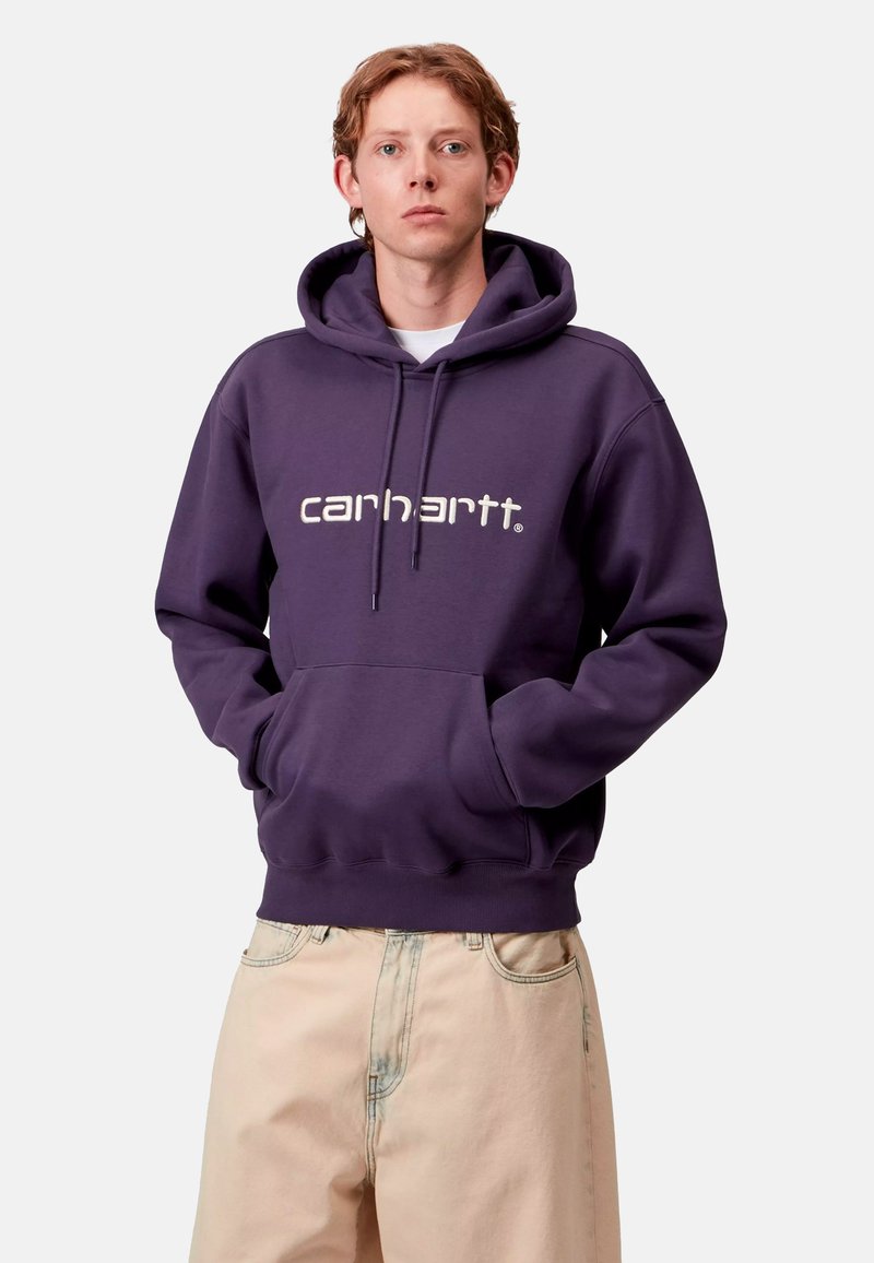 Felpa viola con tasca frontale, polsini e orlo a costine, con il logo "Carhartt" bianco. Abbinata a pantaloni larghi di colore chiaro.