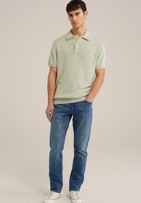 Šviesiai žalia trumpomis rankovėmis polo marškinėliai su lygia apykakle ir trimis mygtukais, derinami su mėlynomis džinsinėmis kelnėmis ir baltomis sportbačiais su raudonais akcentais.