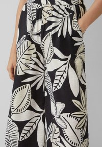 Zwarte stoffen broek met grote witte bladeren en bloemmotieven, elastische tailleband en zijzakken voor extra functionaliteit.