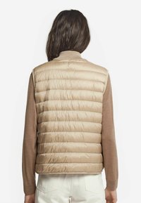Beige puffer väst med horisontell quiltning, ribbad krage och mjuk textur. Bärs över en brun långärmad topp och vita byxor.