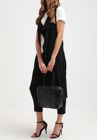 Kvinde iført sort ærmeløs lang vest, hvid T-shirt, sorte cropped bukser, sorte højhælede sandaler, mens hun holder en sort håndtaske i hænderne.