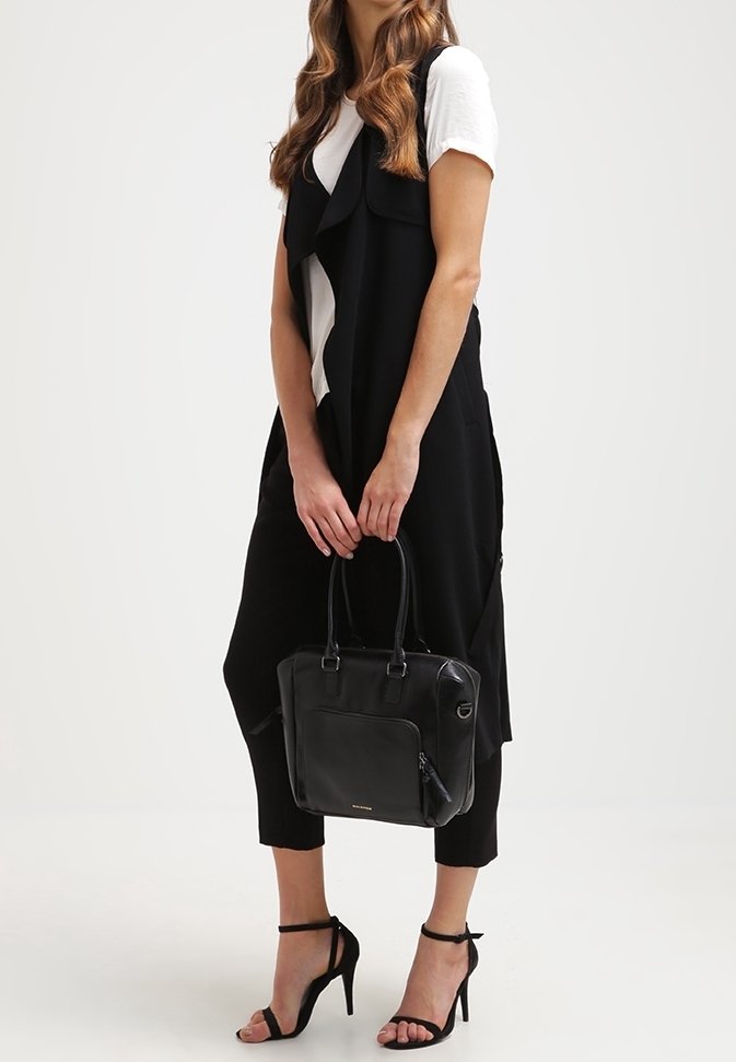 Kvinde iført sort ærmeløs lang vest, hvid T-shirt, sorte cropped bukser, sorte højhælede sandaler, mens hun holder en sort håndtaske i hænderne.
