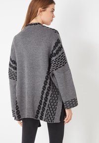 Mujer vistiendo un cárdigan de punto gris con patrones geométricos negros, de espaldas, combinado con pantalones negros, contra un fondo blanco.