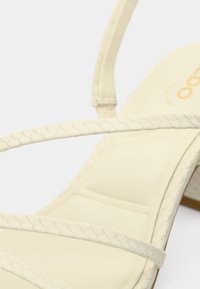 Augsti papēži sandales ar siksniņām bēšā krāsā, ar strukturētām krusteniskām siksnām un polsterētu zolīti ērtībai. Gluda zolītes virsma.