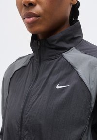 Κοντινό πλάνο ενός ατόμου που φοράει μαύρο και γκρι μπουφάν Nike με φερμουάρ και ψηλό γιακά, εστιάζοντας στο πάνω μέρος του στήθους και στο κάτω μέρος του προσώπου.