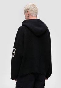 Mustat neulehuppari, jossa on ribbitekstuuri, suuri huppu, löysä istuvuus ja risareuna. Vasemmassa hihassa on näkyvä valkoinen numero "13".