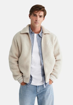 Fleece jacket in off-white with a zipper and collar. Features a textured finish and a subtle logo. Layered over a plaid shirt.