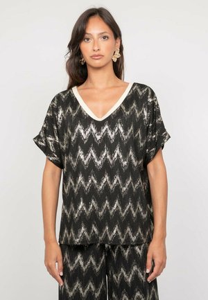 Femme portant un haut à motif zigzag noir et beige avec col en V et un pantalon assorti, accessoirisée de boucles d'oreilles florales beiges, debout devant un fond uni.