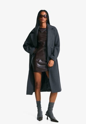 Cappotto grigio di lana con ampi risvolti, chiusura a un bottone; abbinato a un vestito in pelle marrone scuro aderente e calze grigie a coste. Scarpe nere con tacco.