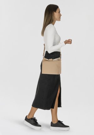 Borsa a tracolla in pelle beige con design strutturato e tracolla regolabile, caratterizzata da una texture semplice e liscia. Indossata con una gonna nera e sneakers.