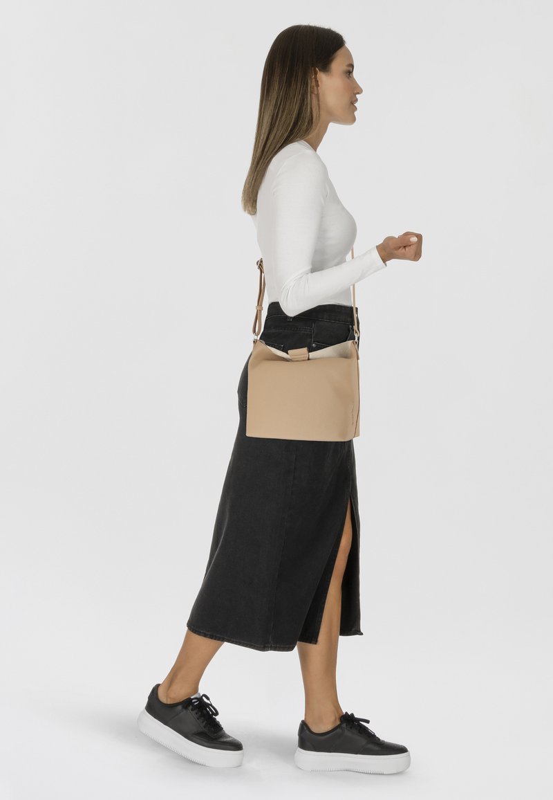Borsa a tracolla in pelle beige con design strutturato e tracolla regolabile, caratterizzata da una texture semplice e liscia. Indossata con una gonna nera e sneakers.