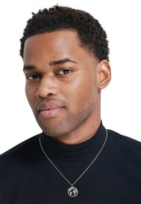 Collier en argent avec un pendentif circulaire affichant un design de carte du monde, porté par-dessus un col roulé noir. Métal lisse et chaîne délicate.