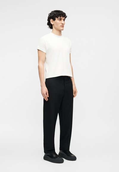 Νεαρός με σγουρά μαλλιά, στέκεται, φοράει απλό λευκό κοντομάνικο πουκάμισο, μαύρο παντελόνι και μαύρα αθλητικά παπούτσια σε λευκό φόντο.