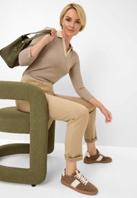 Een ochre gestreepte langemouwen polo, beige slim-fit broeken, bruine suède sneakers met witte strepen en een groene handtas. Ronde groene stoel.
