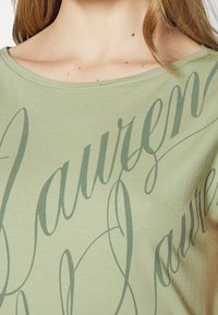 T-shirt en coton vert clair avec un col rond, arborant un grand texte en cursive vert foncé sur le devant, mettant en avant un design fluide.