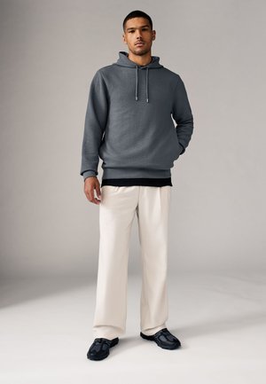 Man staat met een gestructureerde grijze capuchontrui, losse beige broek en zwarte sneakers tegen een effen neutrale achtergrond.