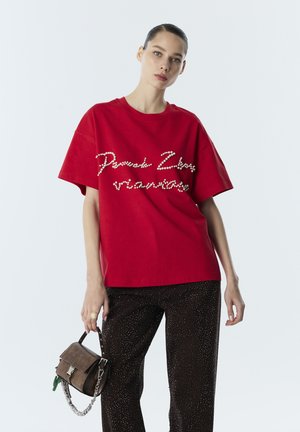Vrouw draagt een rode oversized T-shirt met pareltekst, zwarte glinsterende broek, houdt een bruine handtas met kettingriem vast, staat tegen een witte achtergrond.