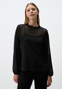 Schwarze transparente Bluse mit gepunkteter Textur, langen Ärmeln, Rundhalsausschnitt und lockerer Passform, über einem einfarbigen schwarzen Unterhemd getragen.