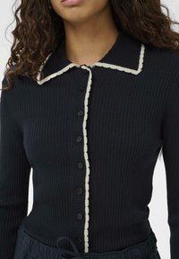 Femeie îmbrăcată cu un cardigan negru dintr-o țesătură ribbord, care se potrivește perfect, având un tiv alb ondulat pe guler și pe linia de nasturi, asortat cu pantaloni negri cu șnur.