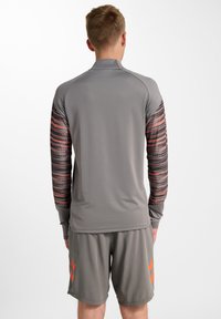 Graues, langärmliges Sporthemd mit gemusterten orangefarbenen und schwarzen Ärmeln, kombiniert mit grauen Shorts mit orangefarbenen Akzenten. Glattes, leichtes Material.