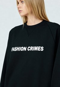 Sweat-shirt noir avec col côtelé et manches raglan, arborant en grande taille le texte blanc audacieux "FASHION CRIMES" sur le devant.