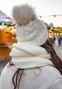 Hvid strikket hue med en fluffy pom-pom på toppen, parret med et tekstureret hvidt tørklæde, der er sikret om halsen, mod en sløret festlig baggrund.