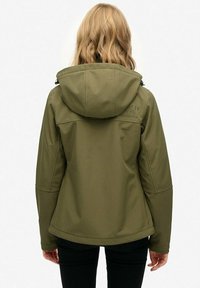 Blondynka w oliwkowej kurtce z kapturem i czarnych spodniach, odwrócona plecami na tle białym.