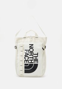 Sac à dos en tissu blanc avec logo et accents noirs. Il dispose d'une poignée supérieure, de bretelles réglables et de sangles latérales verticales.