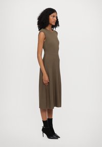 Knee-length olive green dress with ribbed texture, sleeveless design, and fitted bodice; paired with black sock-style heels.