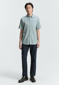 Camisa de manga corta, de color azul claro, con un bolsillo en el pecho, combinada con pantalones azul oscuro y botas negras, de pie contra un fondo liso.