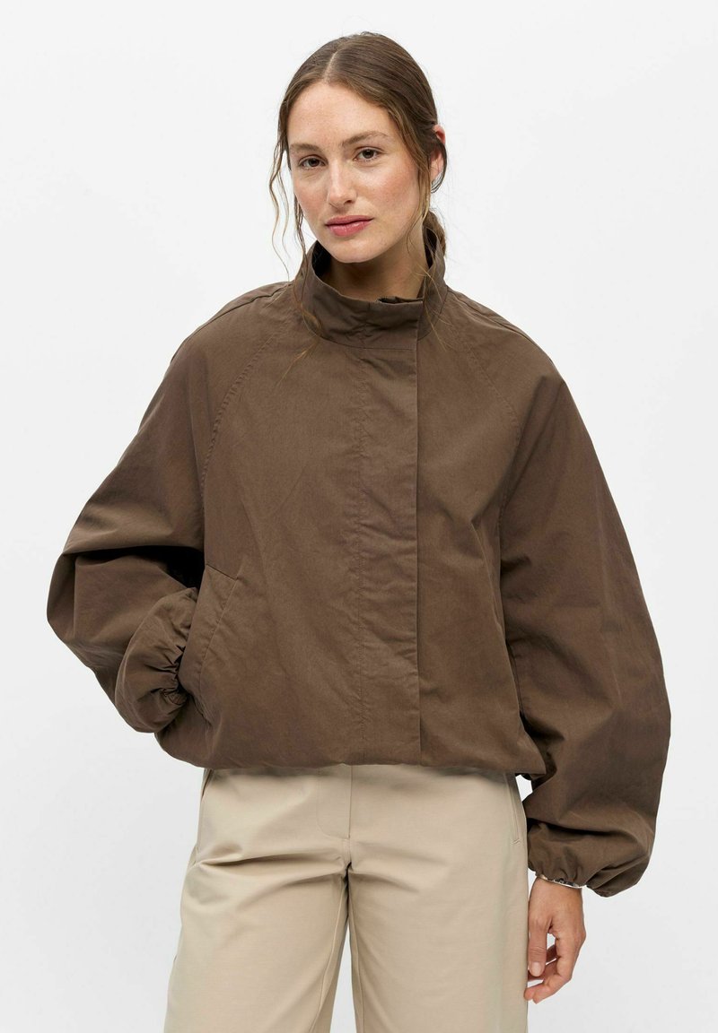 Femme à la peau claire et aux cheveux bruns porte une veste marron oversize à col montant et un pantalon beige, debout devant un fond blanc.